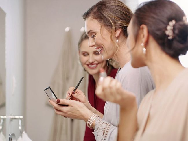 Three people in a salon consult a phone mirror while a makeup artist applies lipstick.