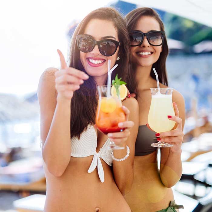 Two women in sunglasses smiling with tropical cocktails at a sunny beach resort.