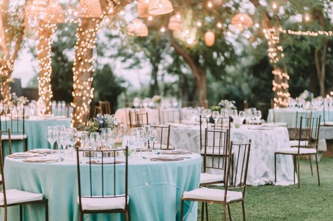 Outdoor reception tables with turquoise linens and string lights among trees at dusk.