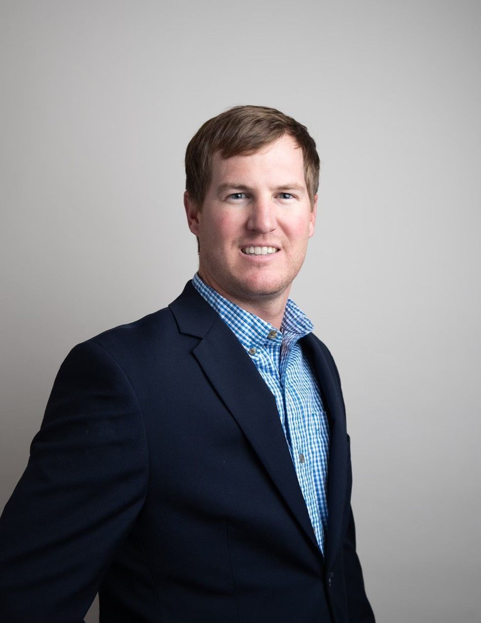 Smiling person in a navy blazer and blue checkered shirt against a light gray background