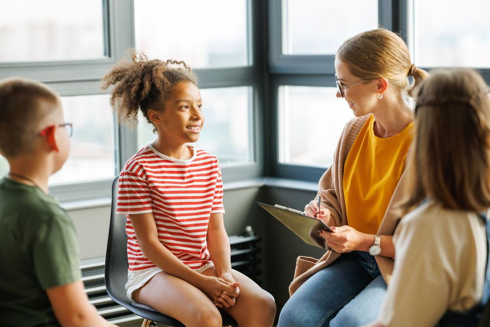 Small group in a bright room talking with a seated facilitator holding a clipboard.