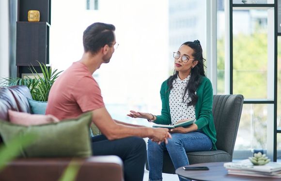 Two people talking on a couch in a bright living room, one holding a notebook.
