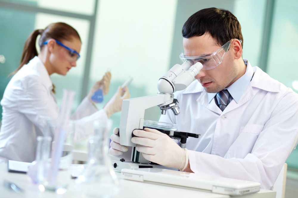 Dos científicos con batas de laboratorio y gafas protectoras trabajando en un laboratorio bien iluminado; uno de ellos utiliza un microscopio.