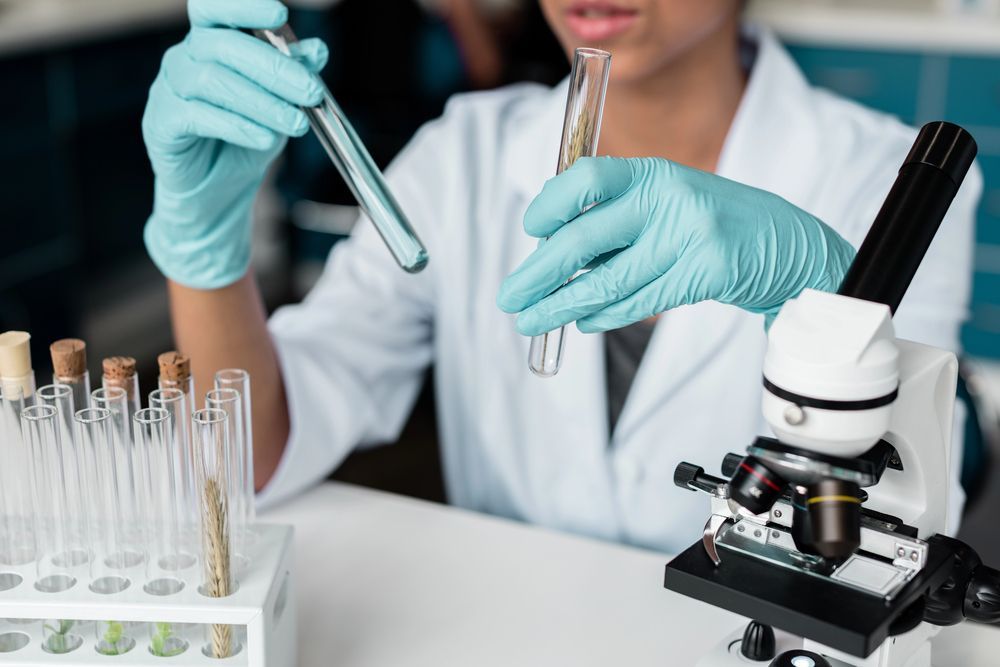 Una persona con bata blanca de laboratorio y guantes azules sostiene tubos de ensayo, con un soporte para tubos de ensayo y un microscopio sobre la mesa.
