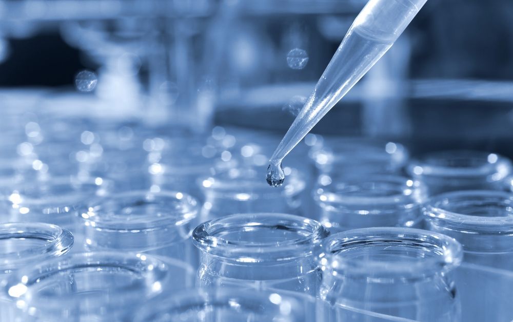 Una pipeta de laboratorio gotea líquido en un vial transparente sobre una bandeja, mientras que otros viales aparecen borrosos al fondo, teñidos de azul.