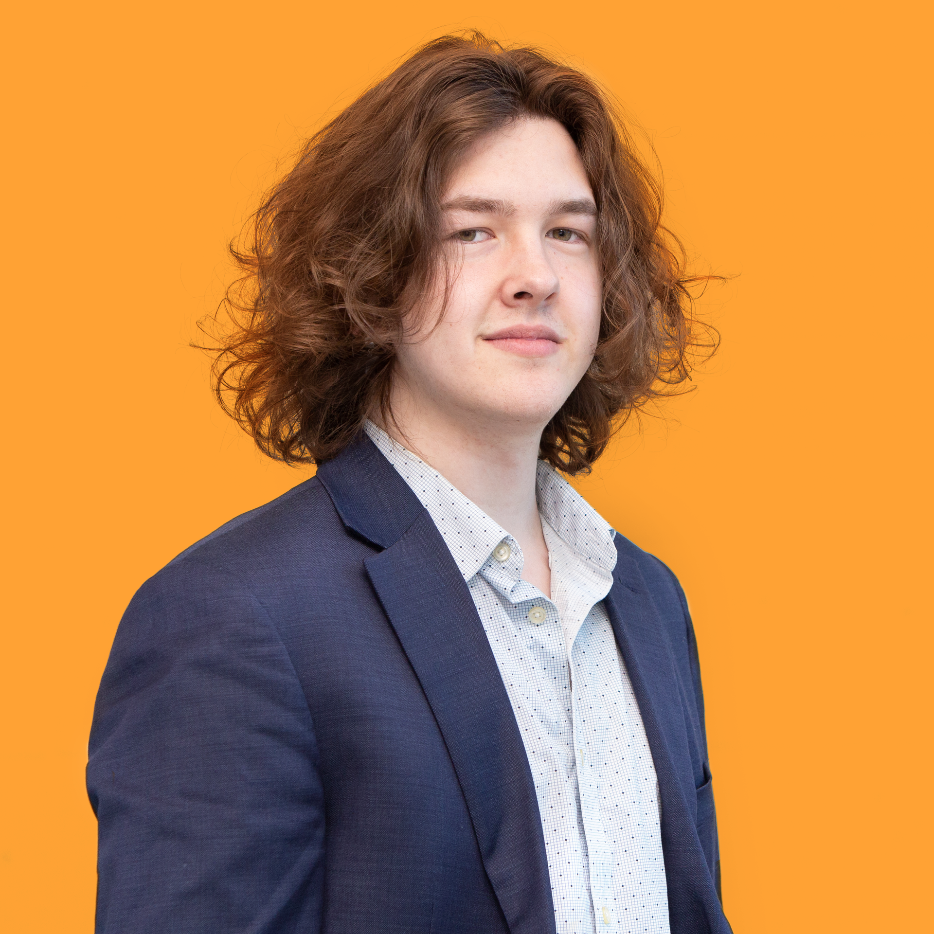 Young man with long brown hair in a blazer and patterned shirt against an orange background.