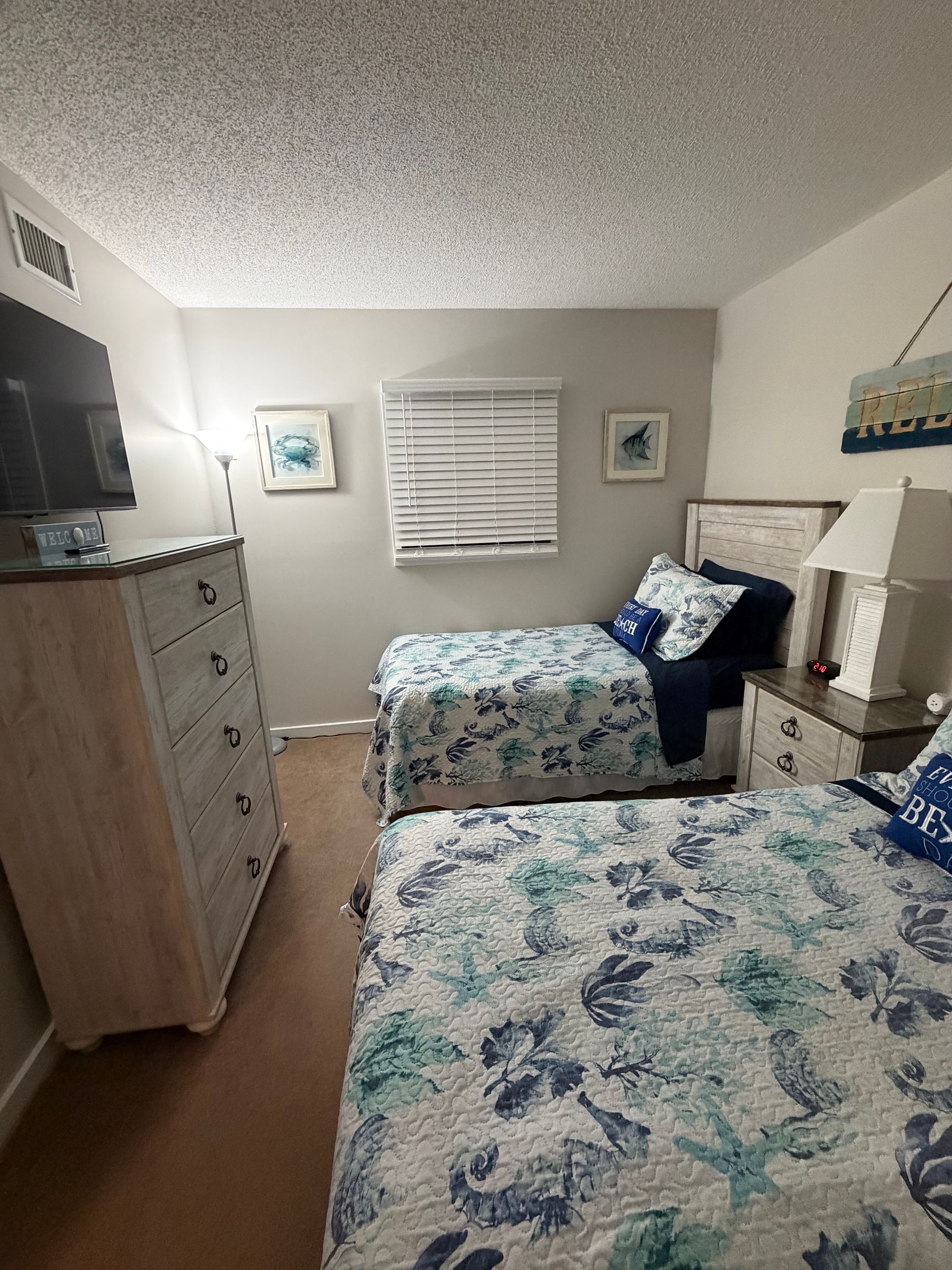 Small bedroom with two twin beds, dresser, TV, and beige walls with blue patterned bedding.