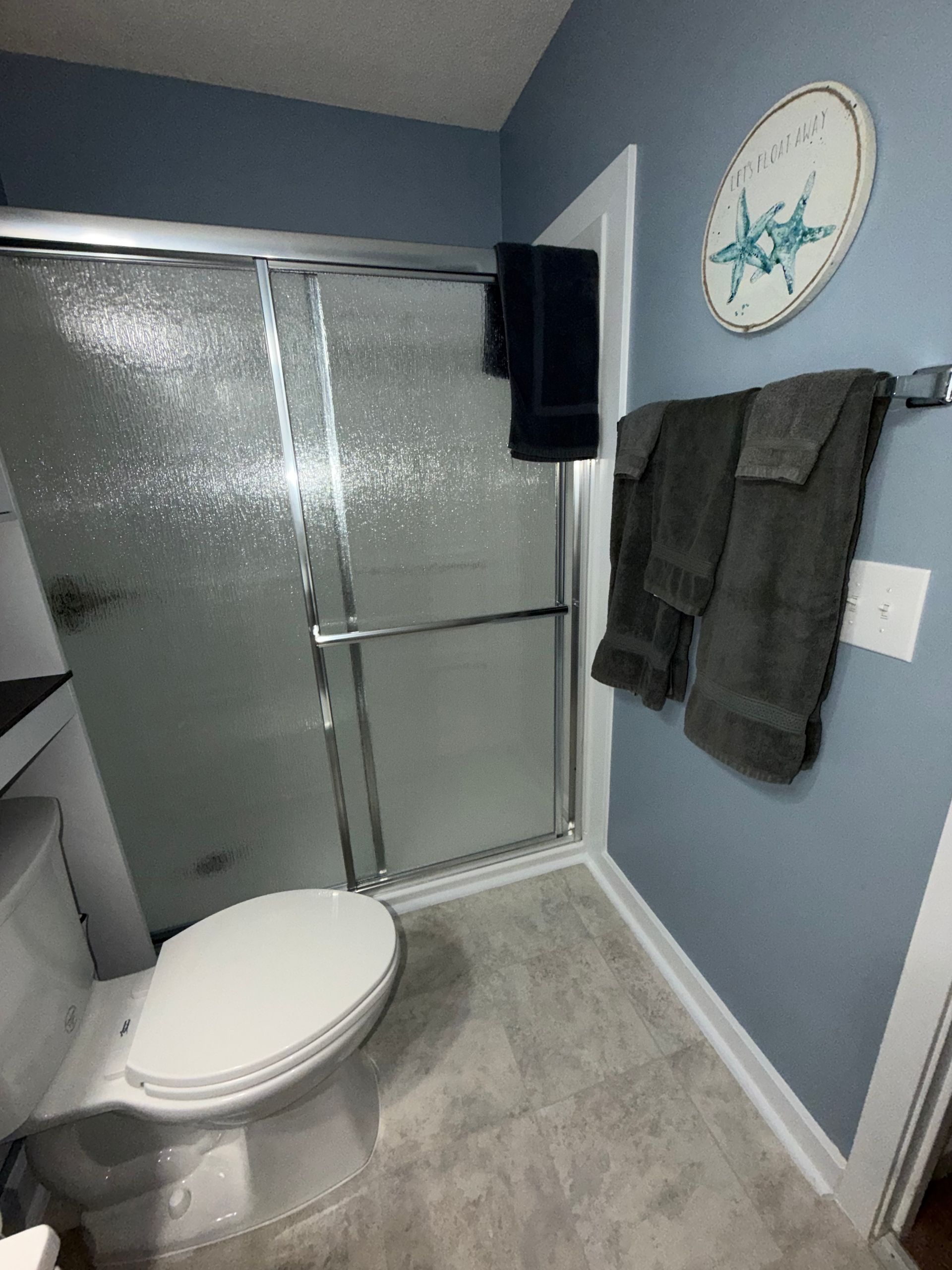 Small bathroom with toilet, frosted shower door, gray walls, and towel rack with dark towels.
