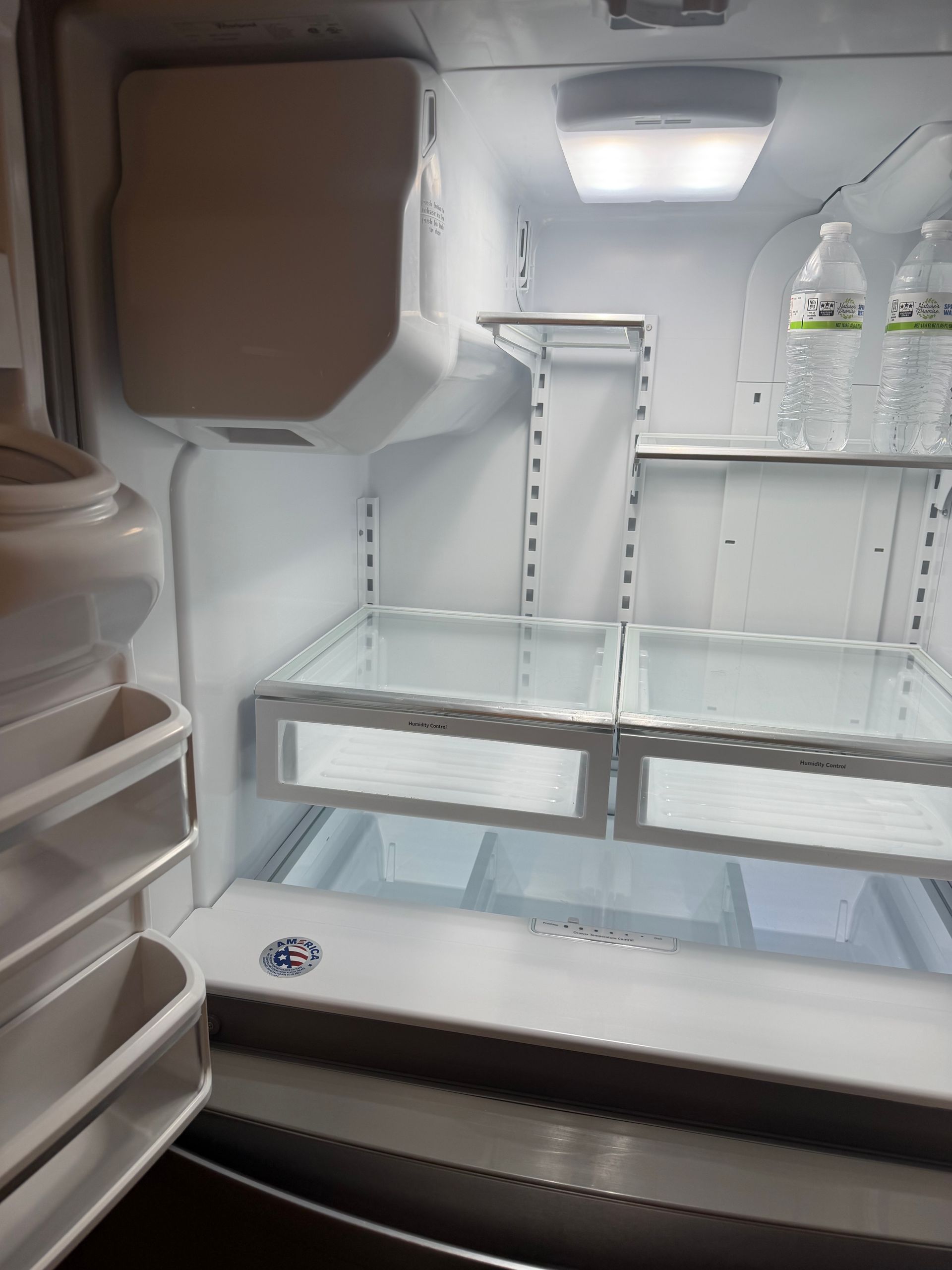 Open refrigerator interior with empty shelves, door bins, and a few bottles on the top right shelf.