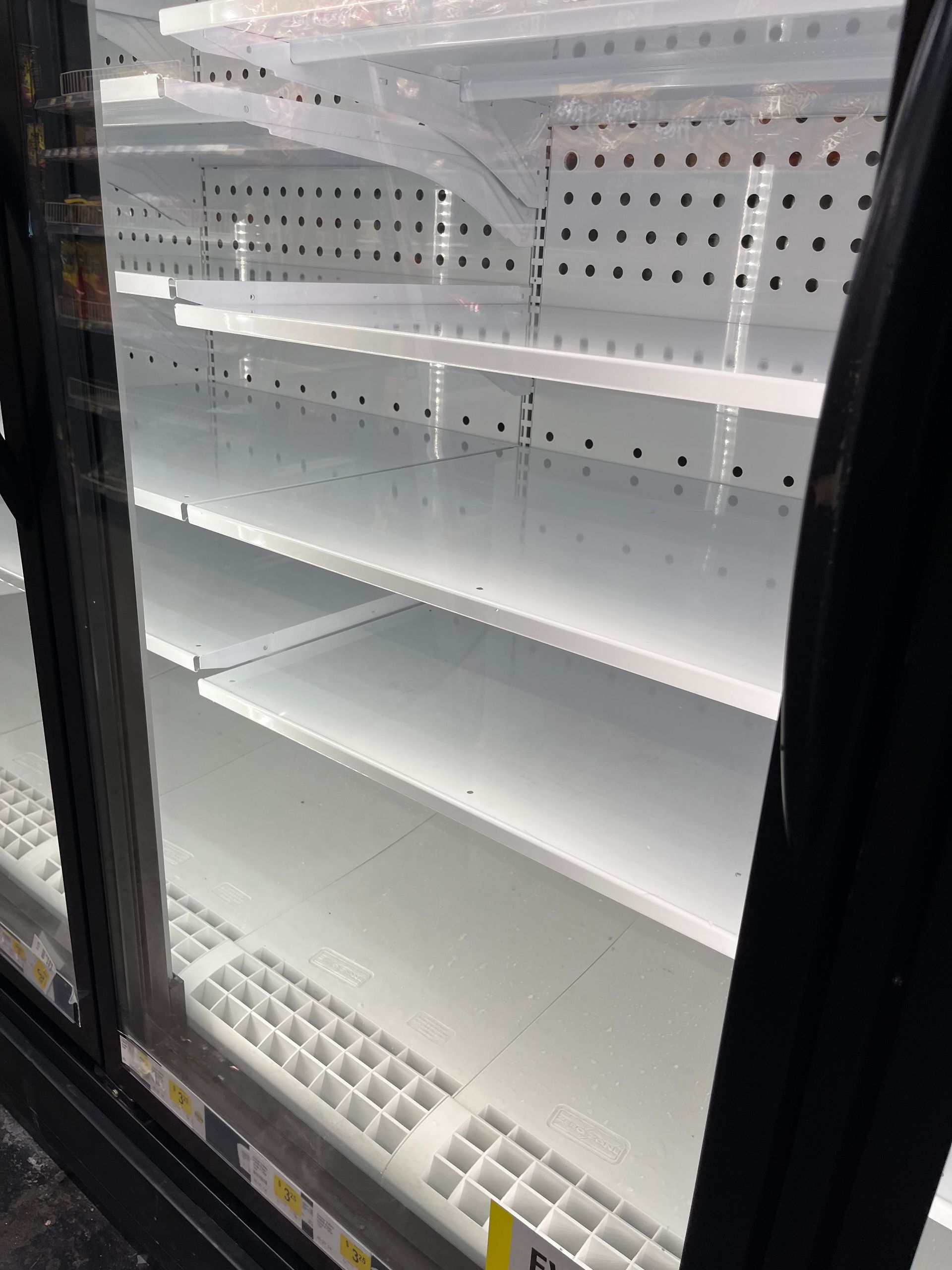 Empty refrigerator shelves in a store, likely due to supply issues or high demand. White shelves are prominently visible.