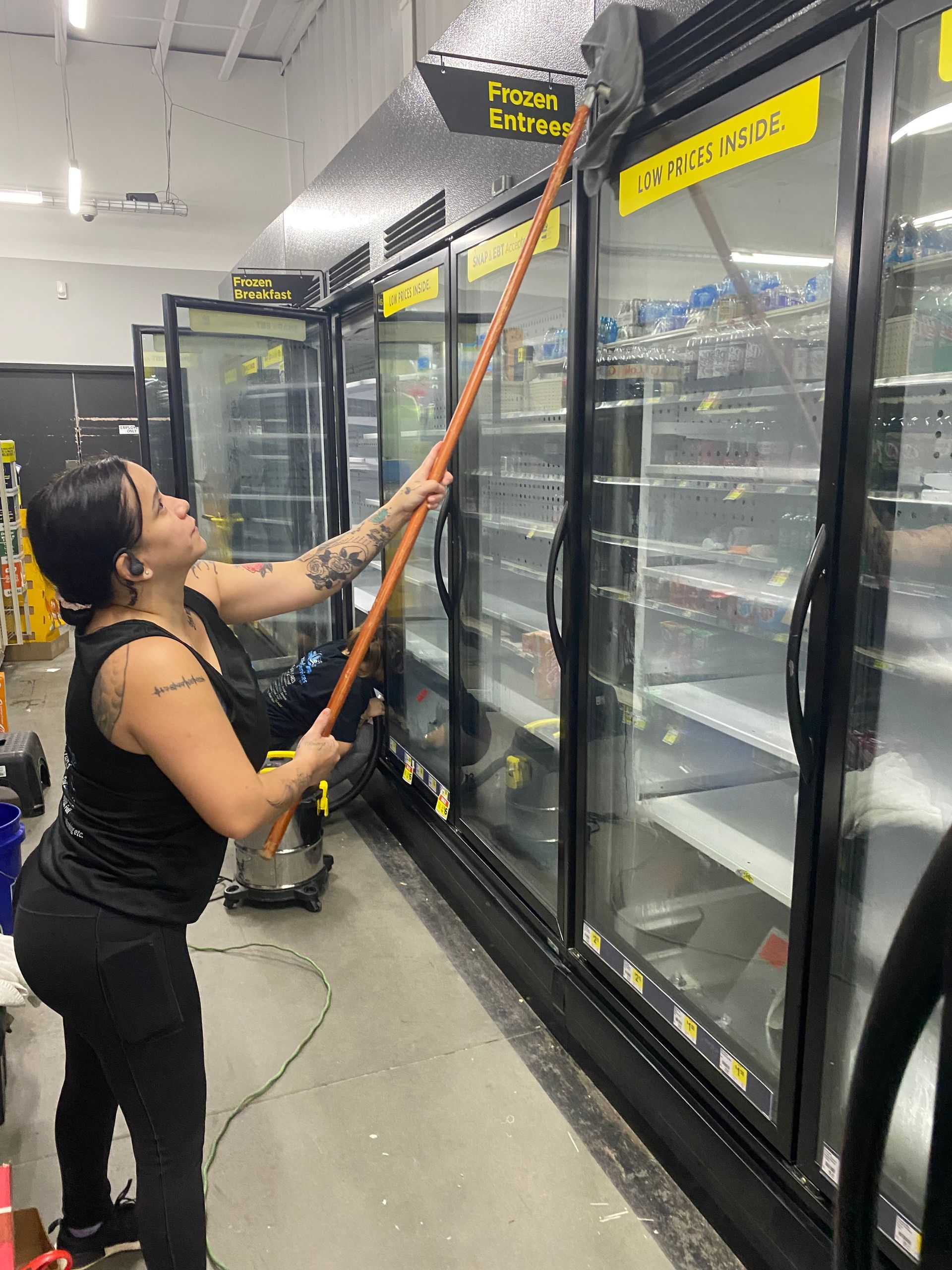 Woman cleaning a grocery store freezer with a long-handled tool. She wears a black tank top and leggings, and has visible tattoos.