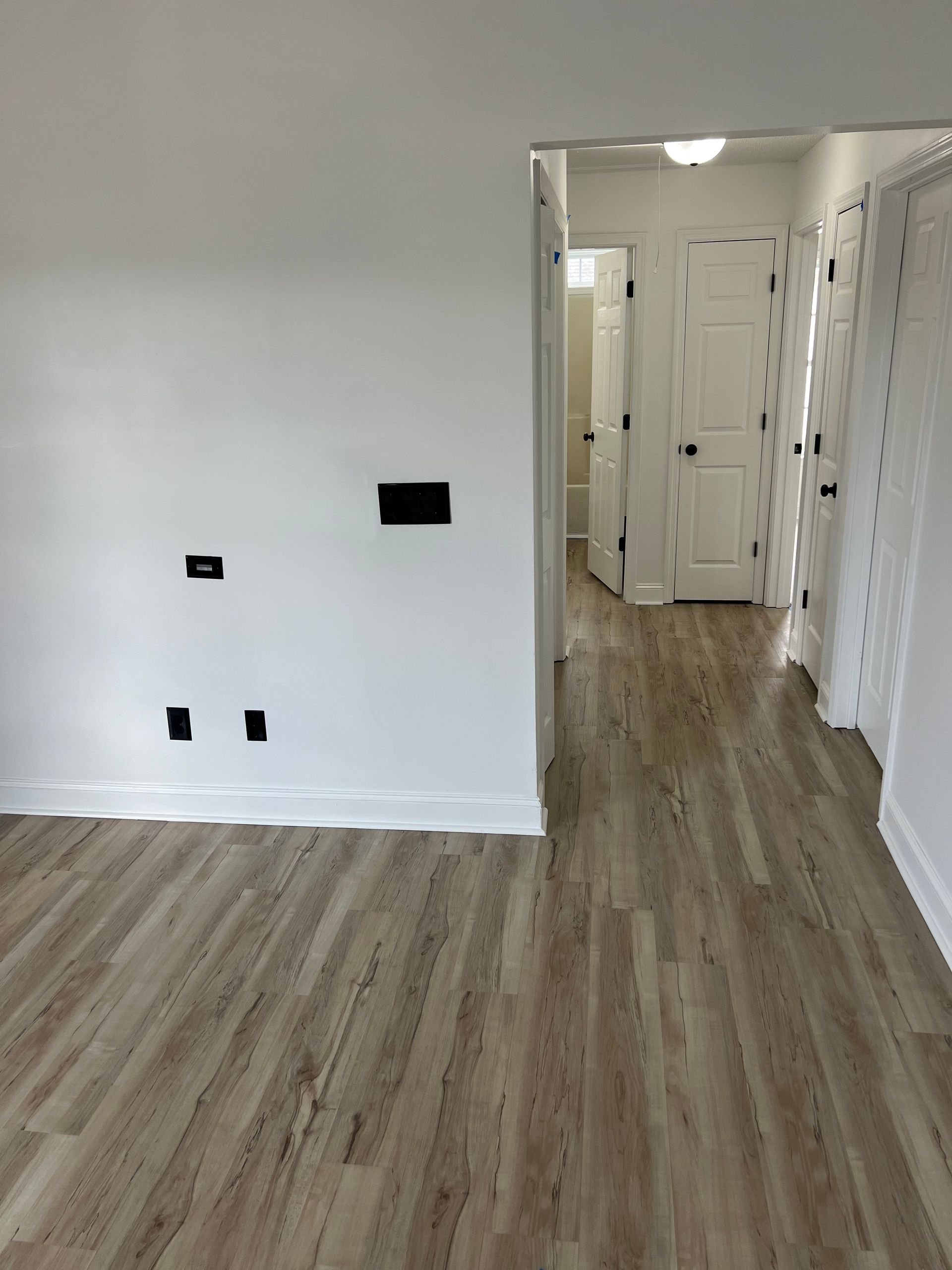 A hallway with wood-look flooring leads to several white doors. A white wall with electrical outlets is on the left.