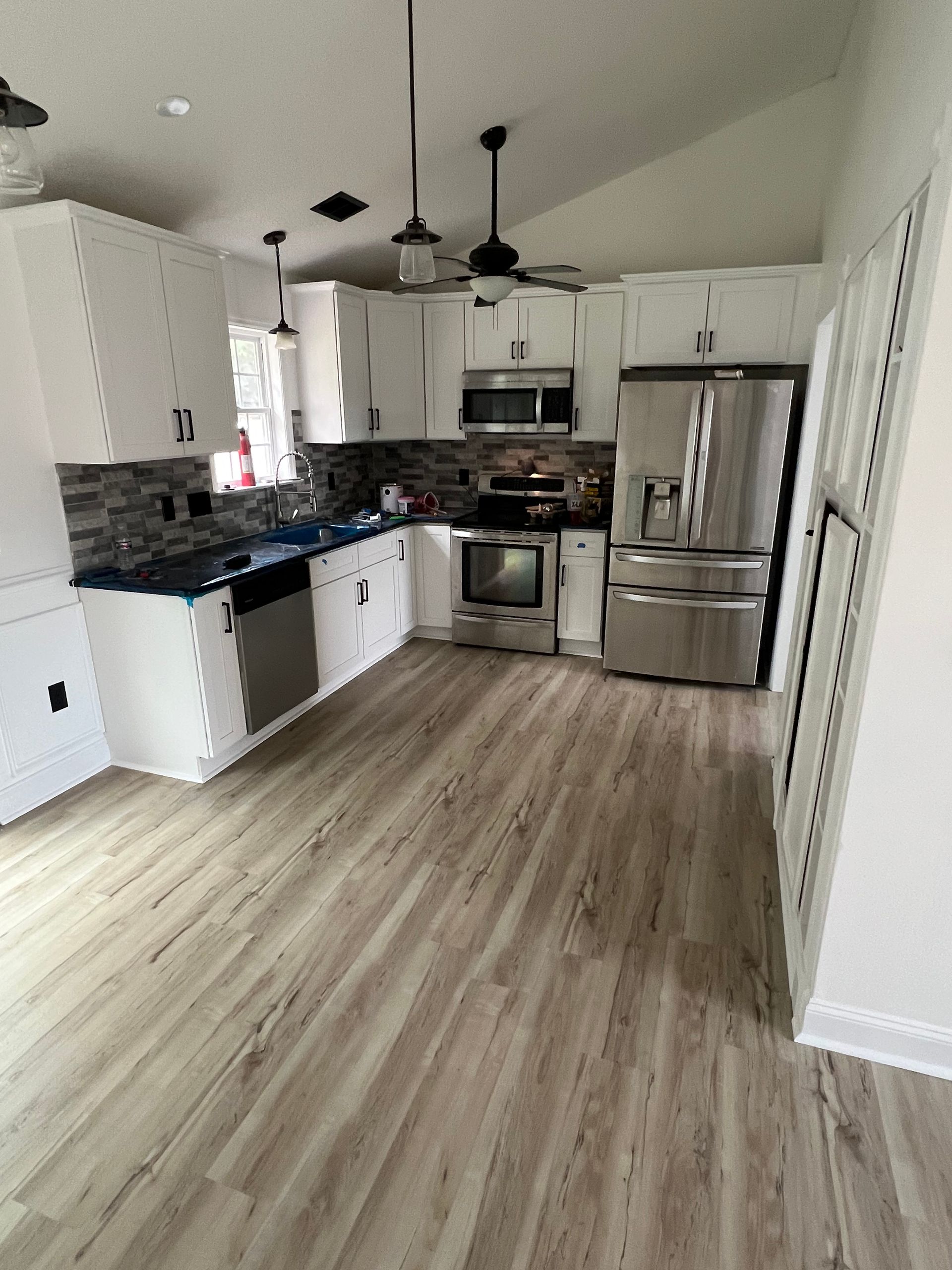 White kitchen with stainless steel appliances, dark countertops, and light wood-look flooring. Black pendant lights hang above the island.