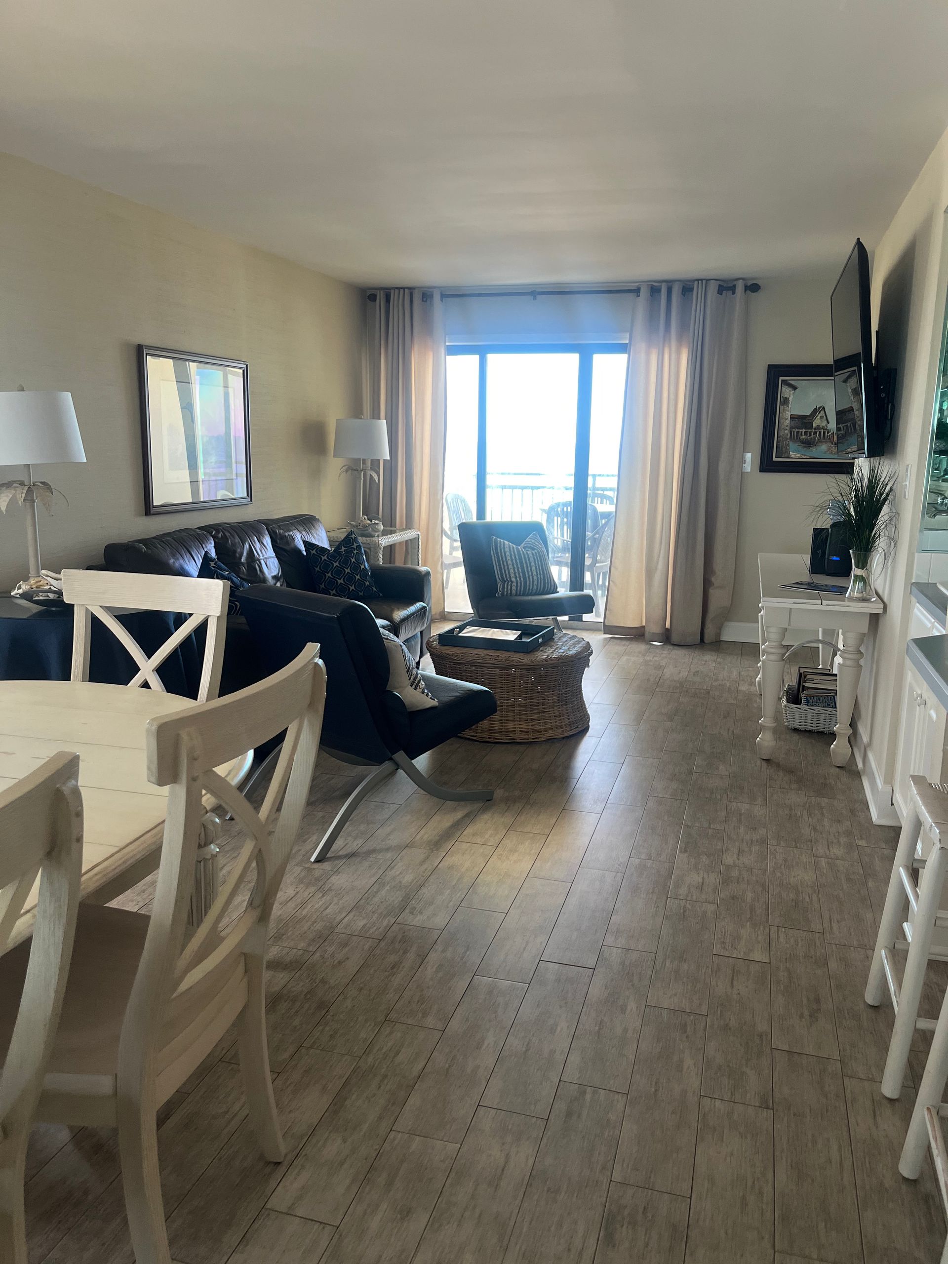 Living room with ocean view: black leather sofa, white dining chairs, and a circular wicker coffee table. Beige curtains frame a sliding glass door.