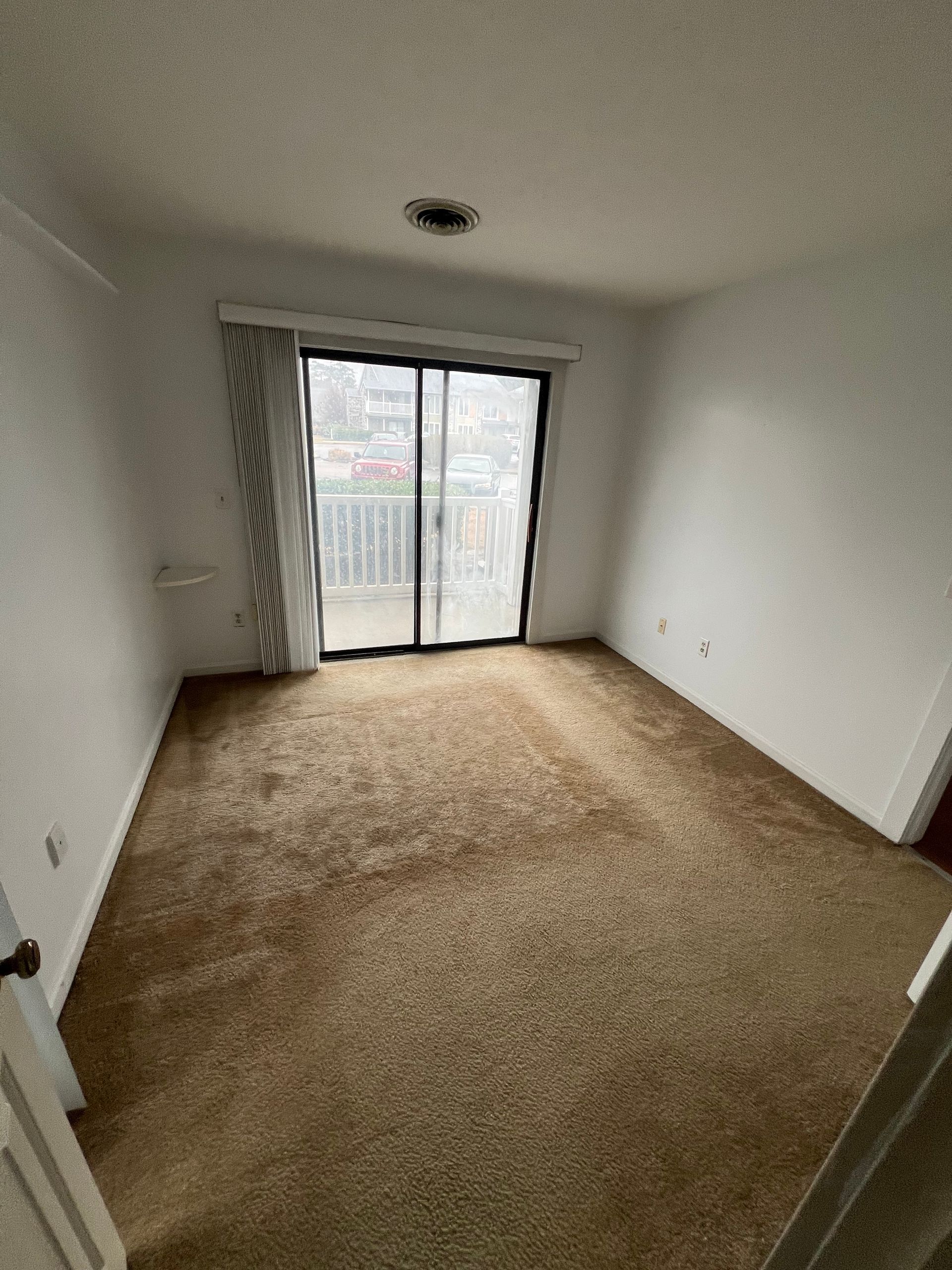 Empty bedroom with brown carpet, sliding glass door leading to a balcony, and white walls.