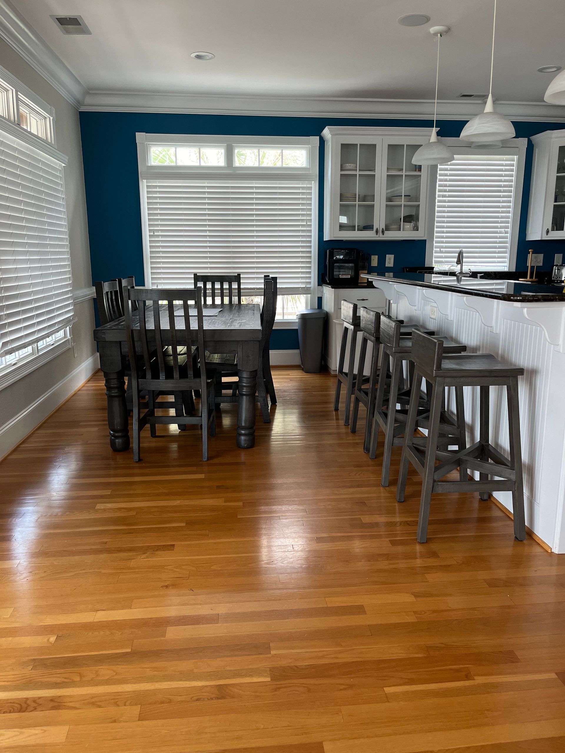 Wooden floor and gray furniture in a dining area. Includes a table and chairs, kitchen island with stools, and window with blinds.