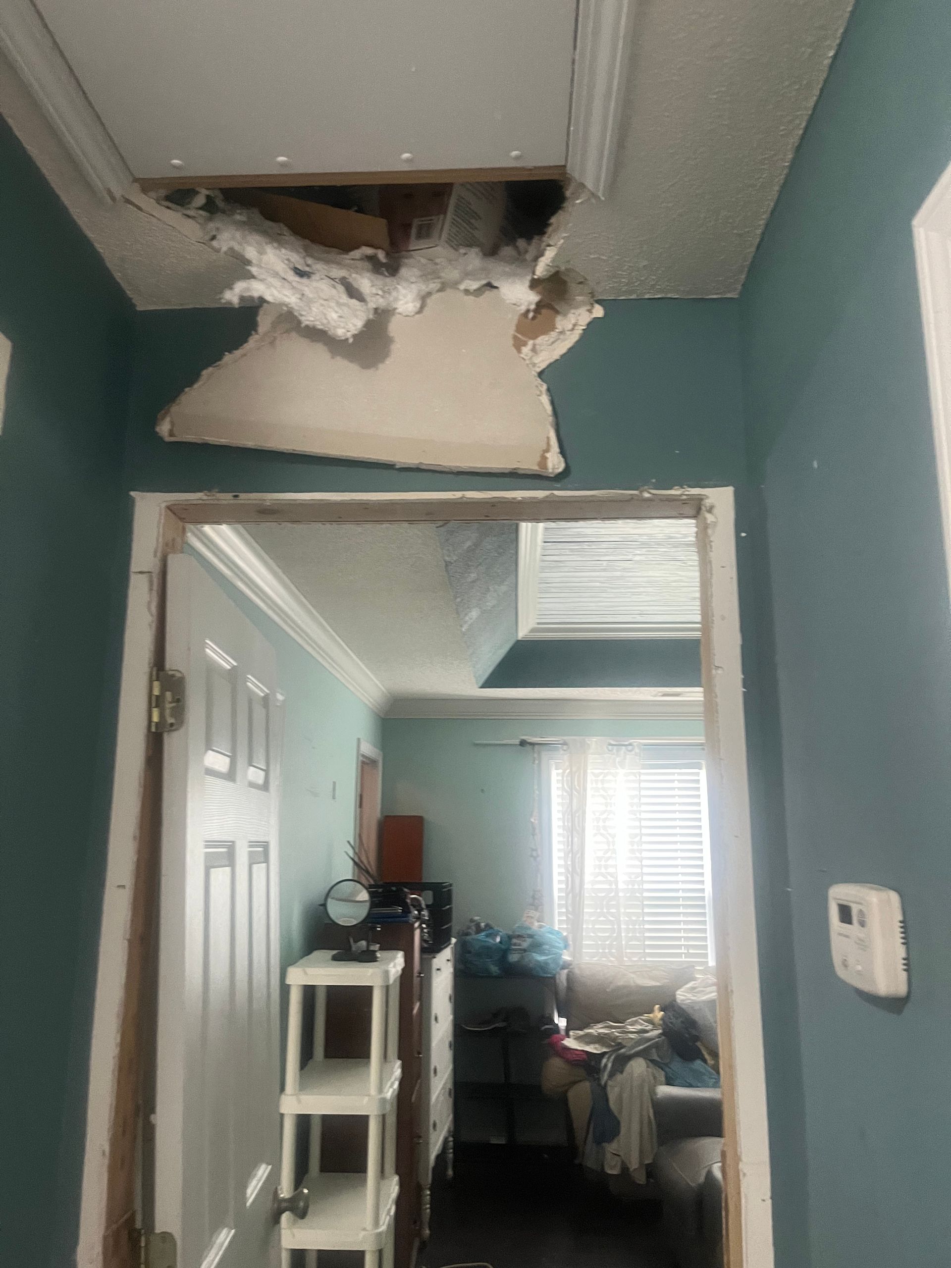 Hallway with damaged ceiling; hole in the ceiling revealing insulation. Doorway leads to a room with a window.