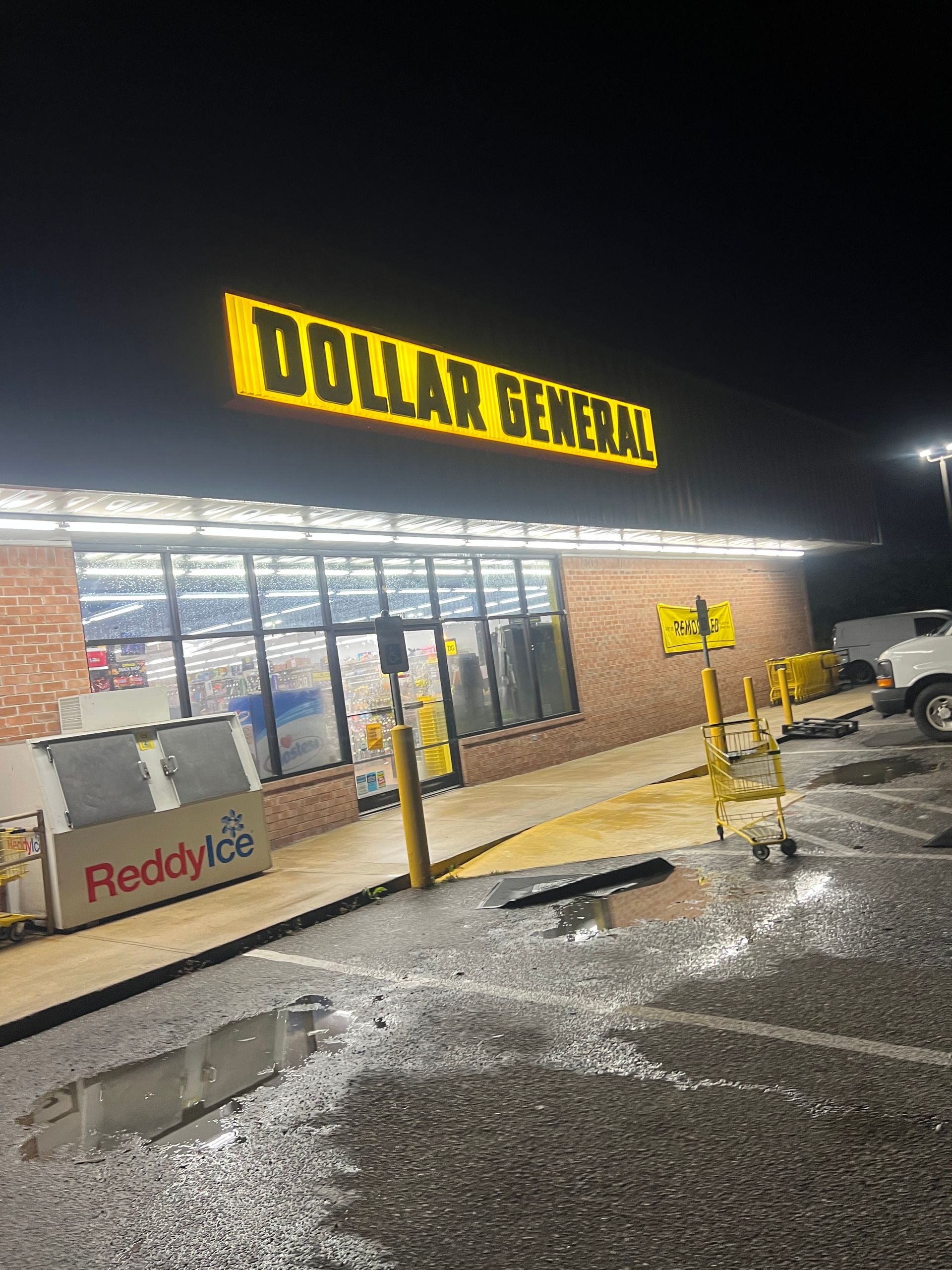 Dollar General store exterior at night with illuminated sign, brick facade, and wet parking lot. A Reddy Ice cart is visible outside.