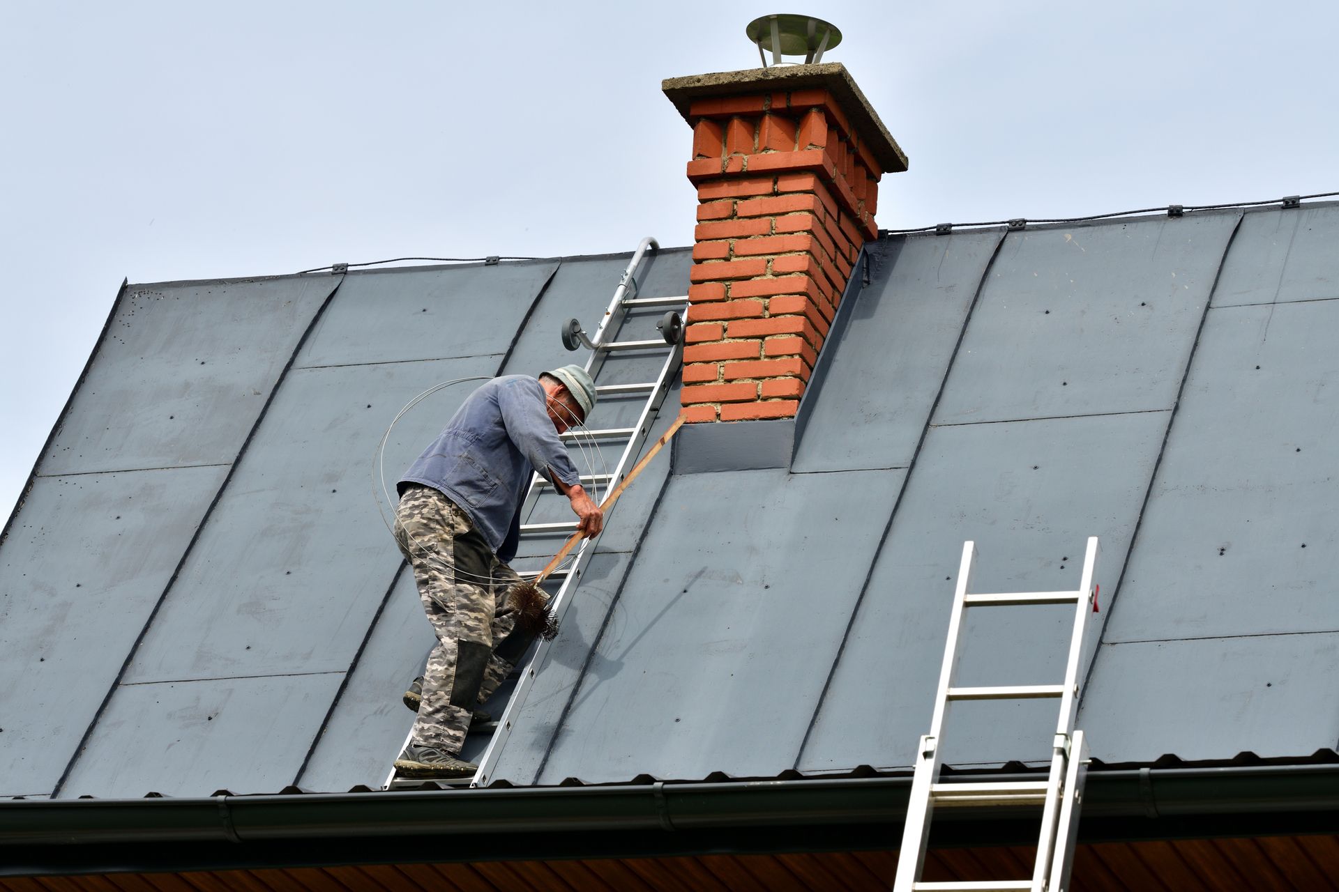 Man on a roof climbs a ladder to a chimney. Gray roof, brick chimney, clear sky. Man on a roof climbs a ladder to a chimney. Gray roof, brick chimney, clear sky.