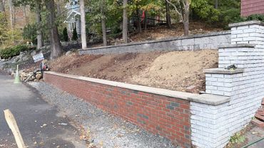 Stone retaining wall and paved area, with trees and fallen leaves in the background.