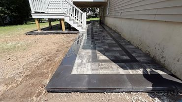 Workers laying pavers on a gravel bed, outdoors. One kneels, placing a paver; two others stand nearby.