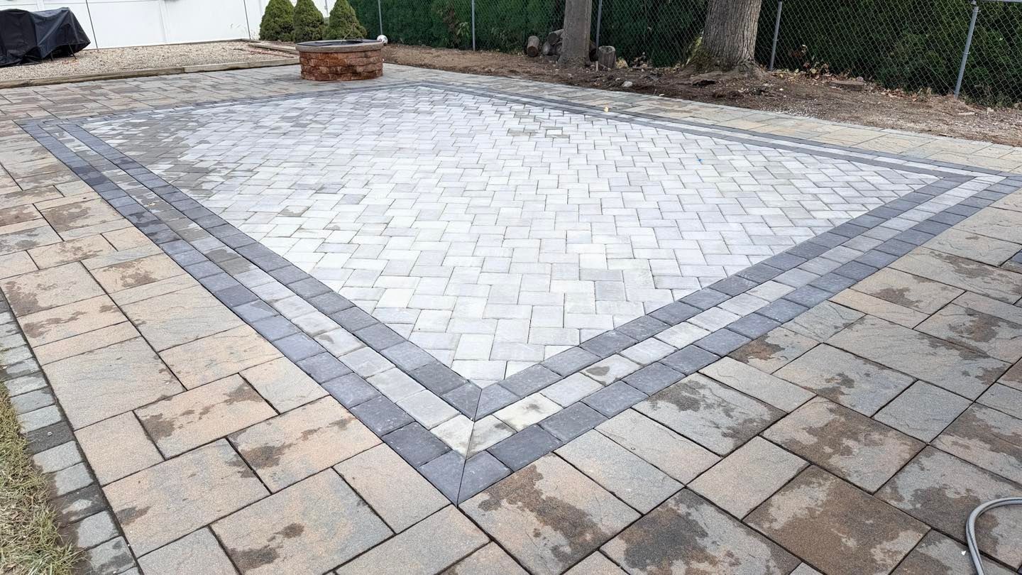 Person in red gloves laying grey pavers on a prepared sand base, outdoors. A rectangular patio made of light gray herringbone pavers with a dark gray border, set in an outdoor yard.