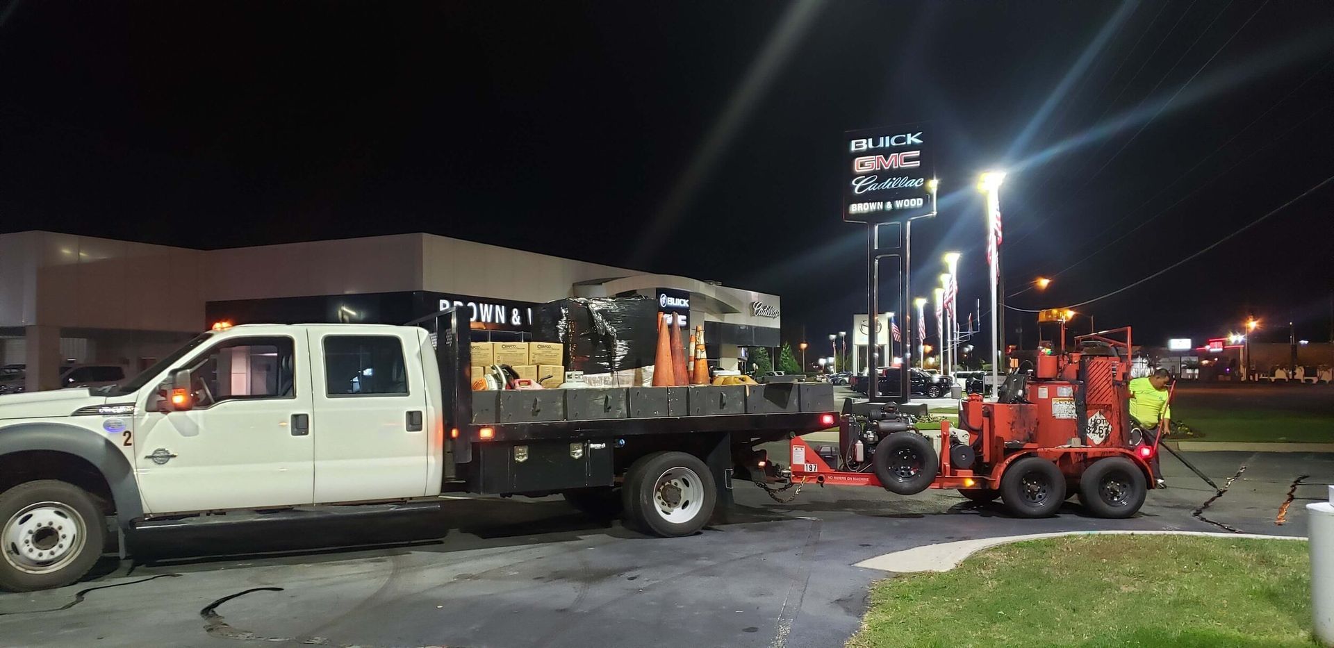 A white truck is towing a red forklift in a parking lot at night.