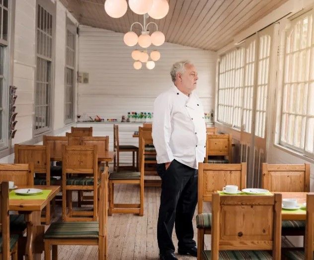 Chef in white uniform looks out the window of an empty, light-filled restaurant with wooden tables and chairs.