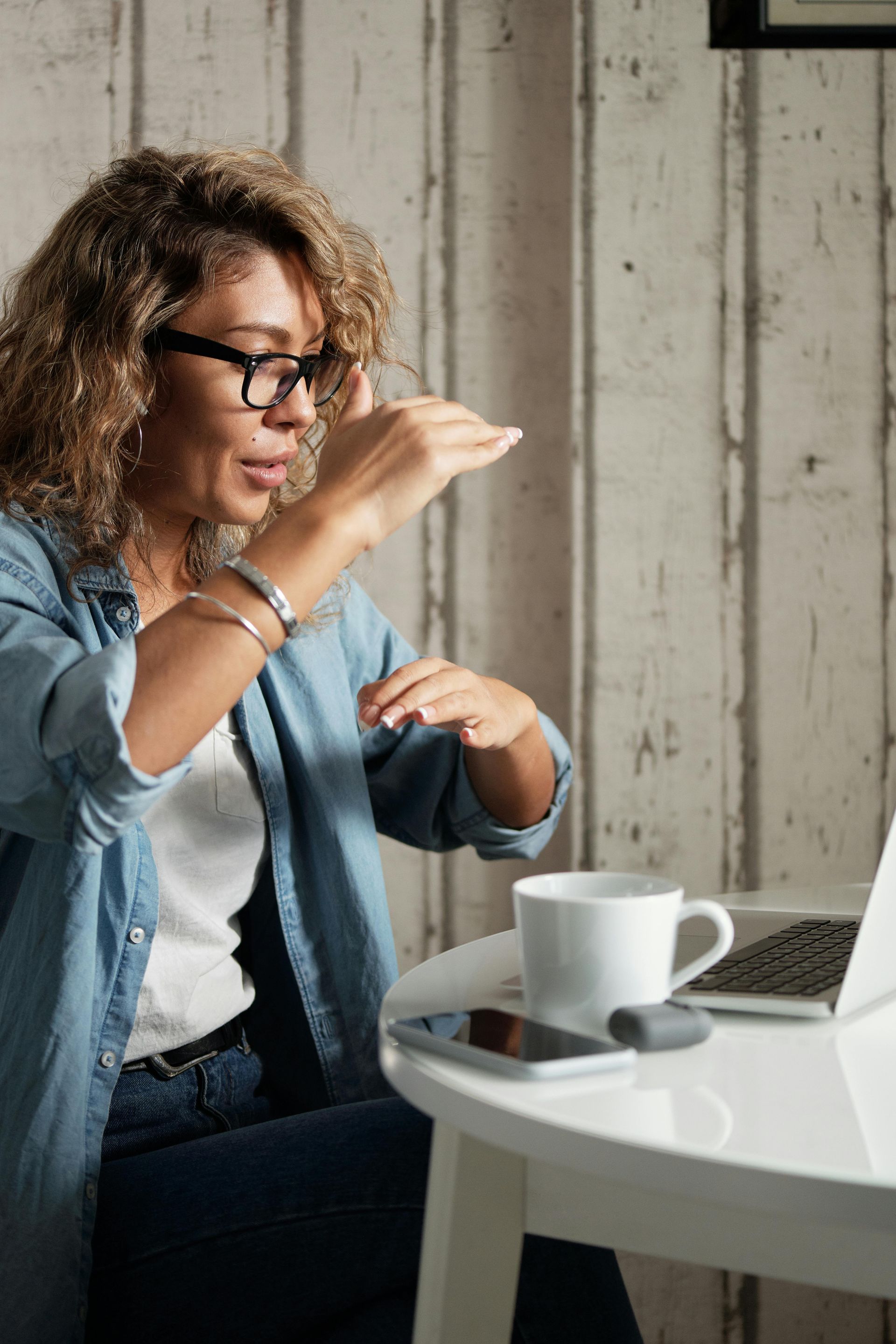 Woman with curly hair and glasses gesturing while on a video call with a laptop and coffee cup at a white table.