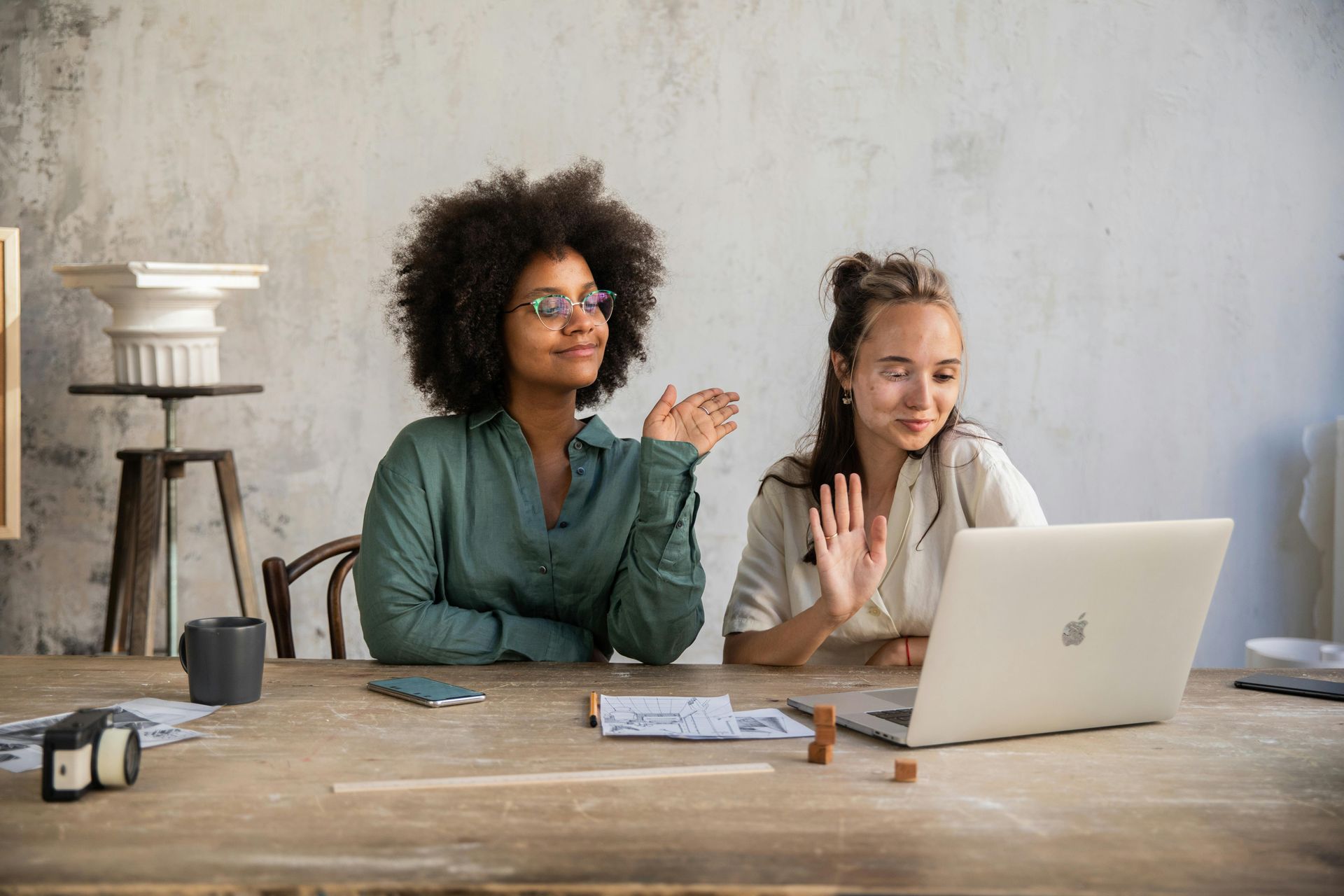 Two women wave at a laptop screen at a wooden table in a connected momentum session. One with curly dark hair wears glasses, and the other has long, light hair.