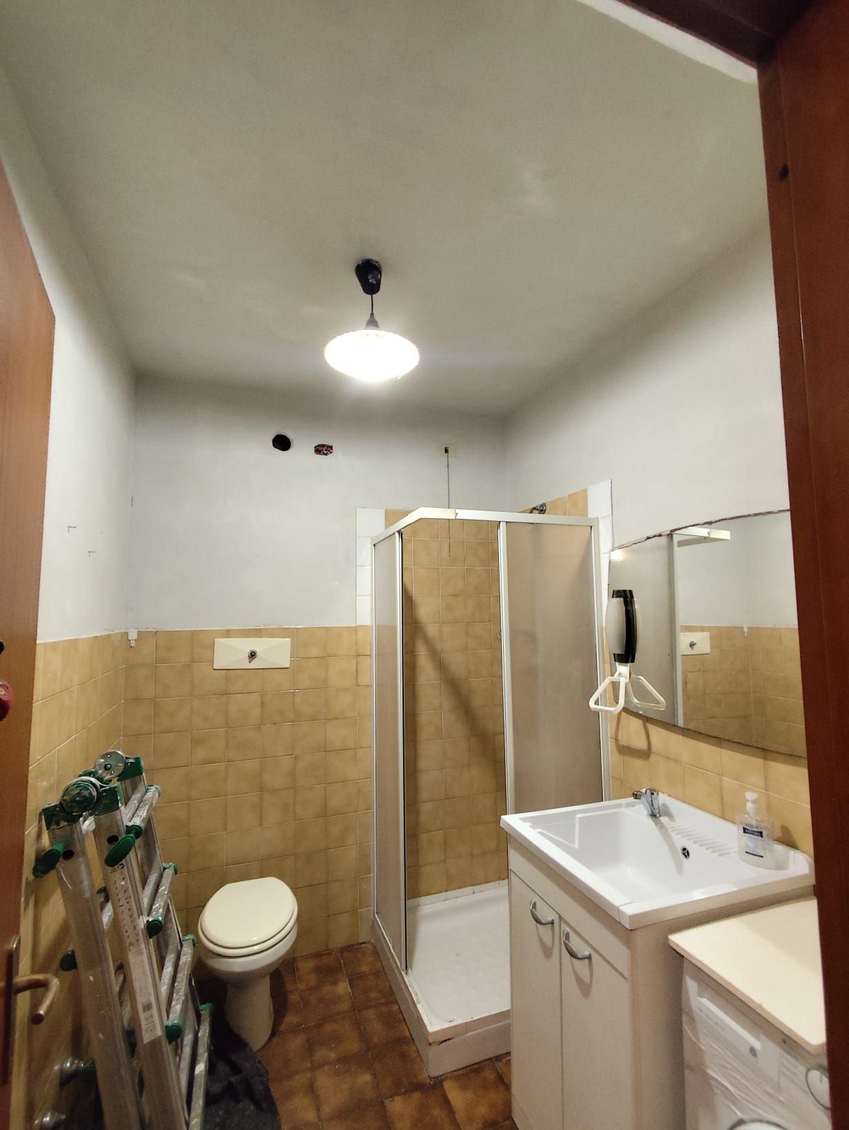 Piccolo bagno con box doccia, lavandino, WC, piastrelle beige e specchio.
