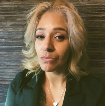 A person with wavy, shoulder-length blonde hair, wearing a dark blazer and a heart-shaped necklace against a dark background.