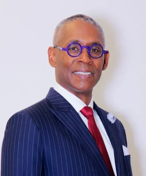 A man in a navy pinstripe suit, white shirt, red tie, and purple-rimmed glasses smiling against a plain white background.