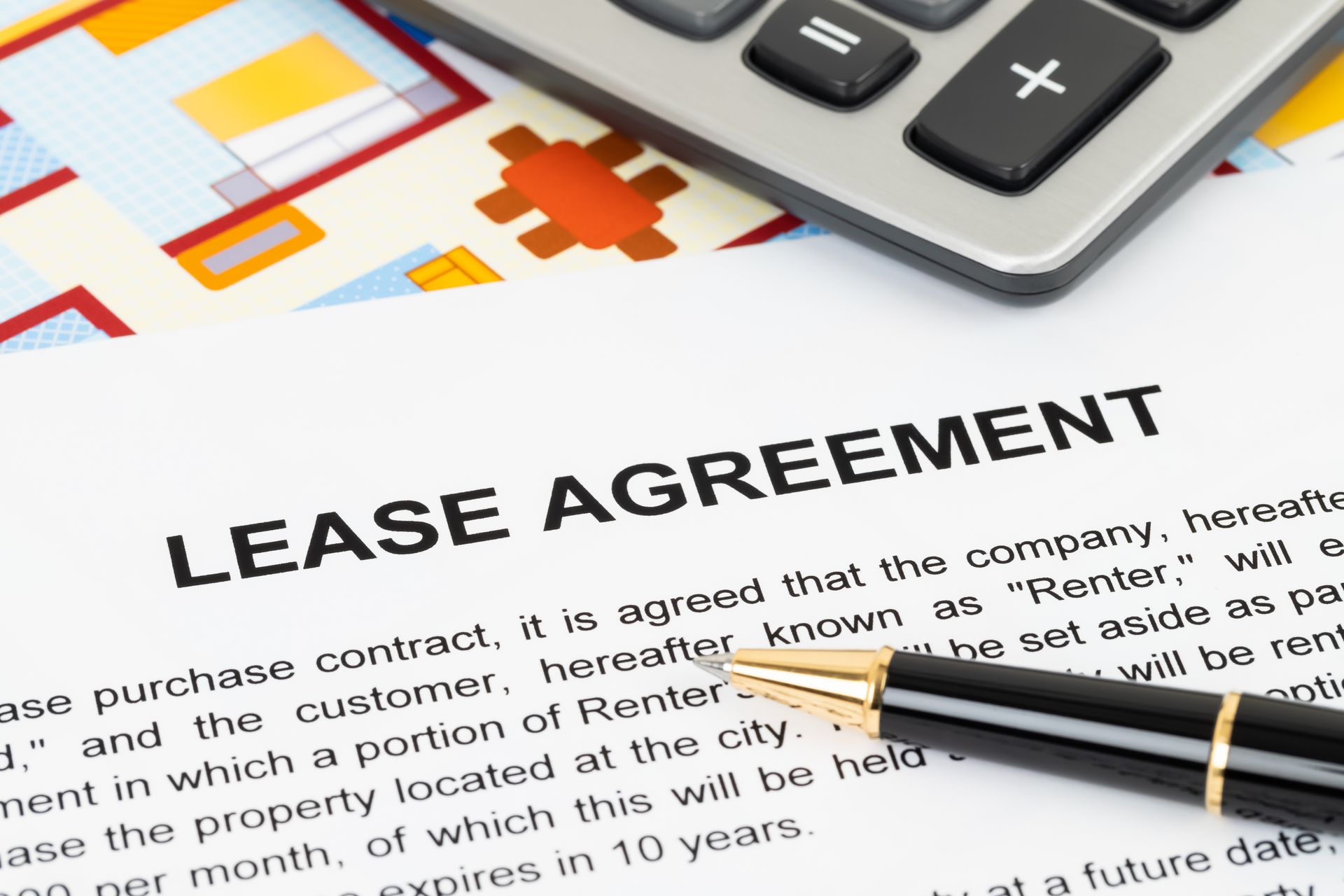 A fountain pen rests on a paper lease agreement, with a calculator partially visible on a floor plan in the background.