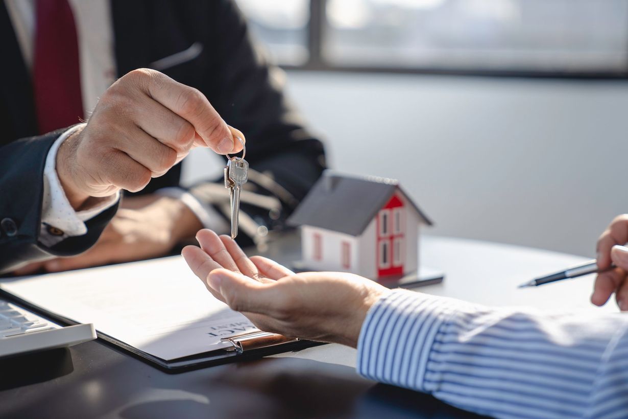A person in a suit hands keys to another person over a table featuring a small model house and documents.