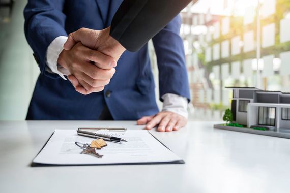 Two people in business suits shake hands over a contract with keys, beside a miniature house model on a desk.