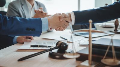 Two people in business suits shake hands over a legal contract, with a judge’s gavel and scales of justice on the table.