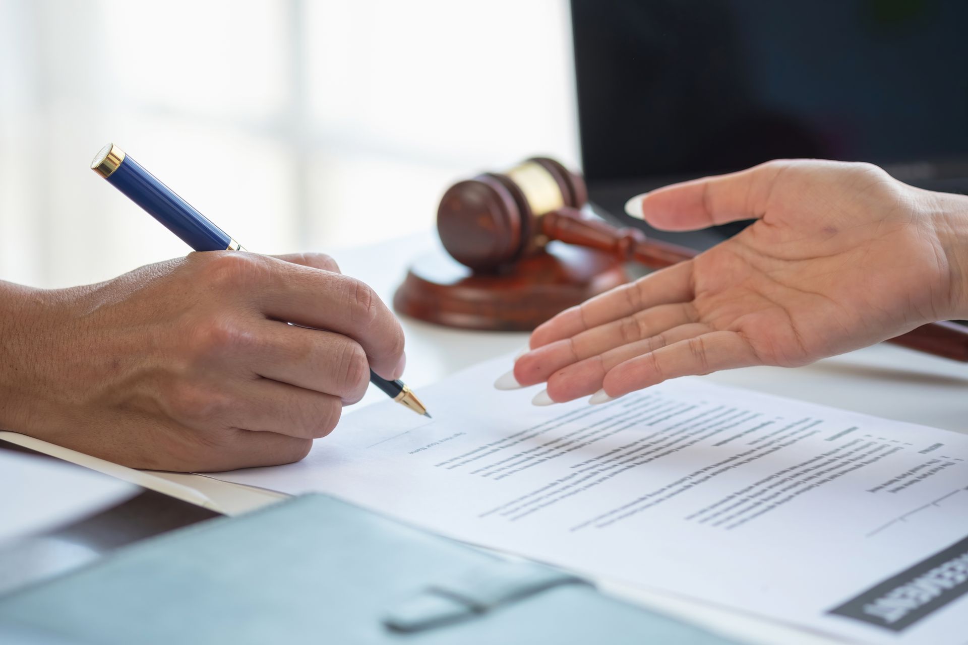 A person signs a legal document with a pen while another person gestures toward the signature line near a wooden gavel.