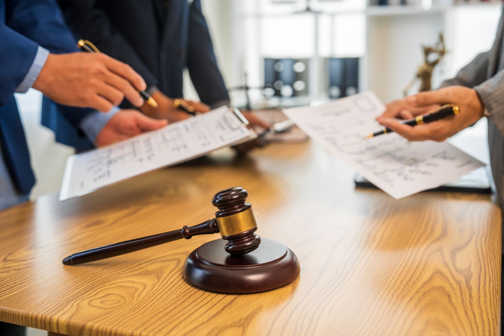 A wooden judge's gavel rests on a desk in the foreground, with people reviewing legal documents in the background.