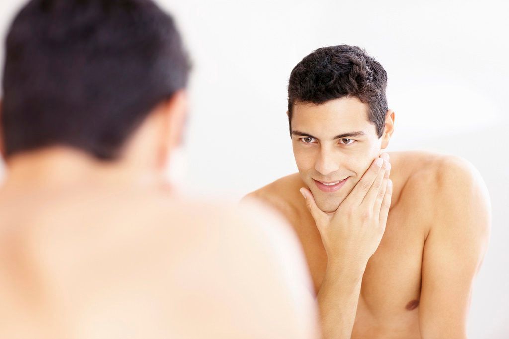 Man looking at his reflection in a bathroom mirror, touching his face with a slight smile