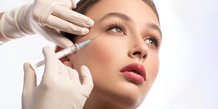 Cosmetic injector preparing a facial syringe for a woman with closed eyes in a bright clinic setting