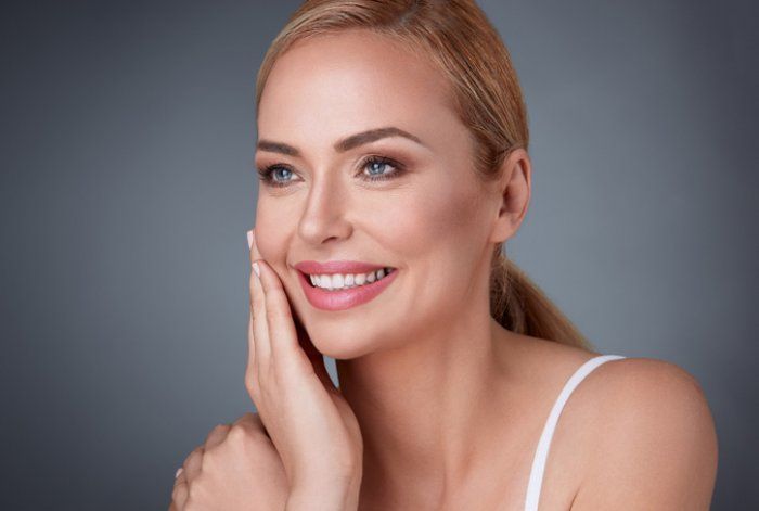Smiling woman with blonde hair touching her cheek against a gray background