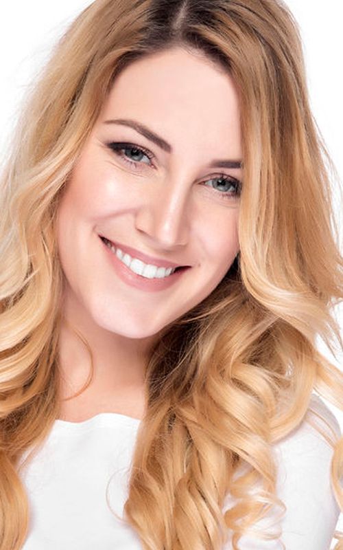 Smiling blonde woman with wavy hair in a white top, softly lit against a plain background