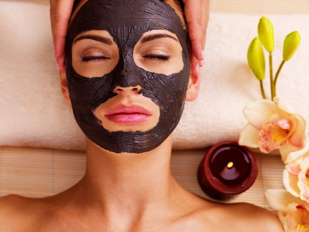 Woman with a black facial mask lying on a spa towel beside flowers and a candle