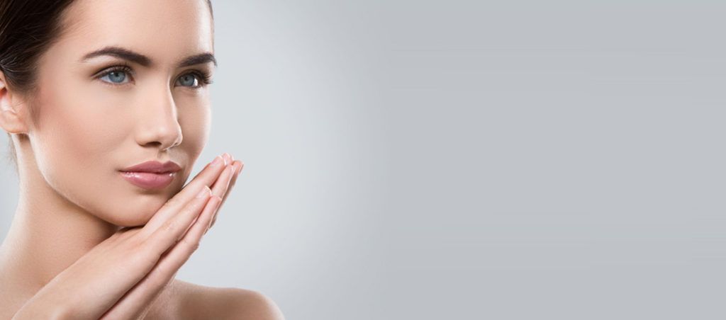 Close-up of a woman with clear skin resting her hands on her face against a gray background