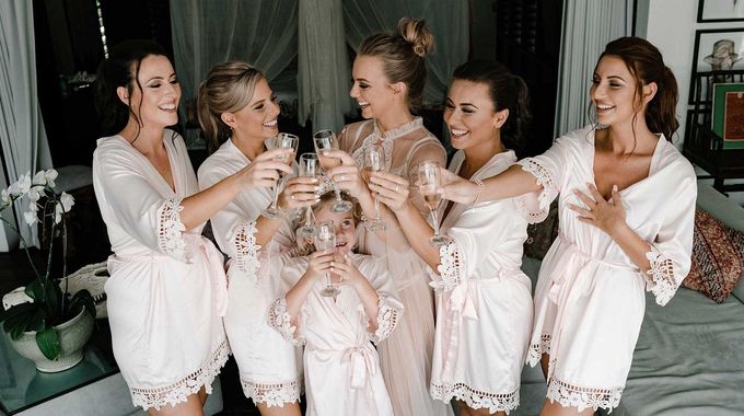 Five people in white robes toast champagne together in a bright bedroom before getting ready.