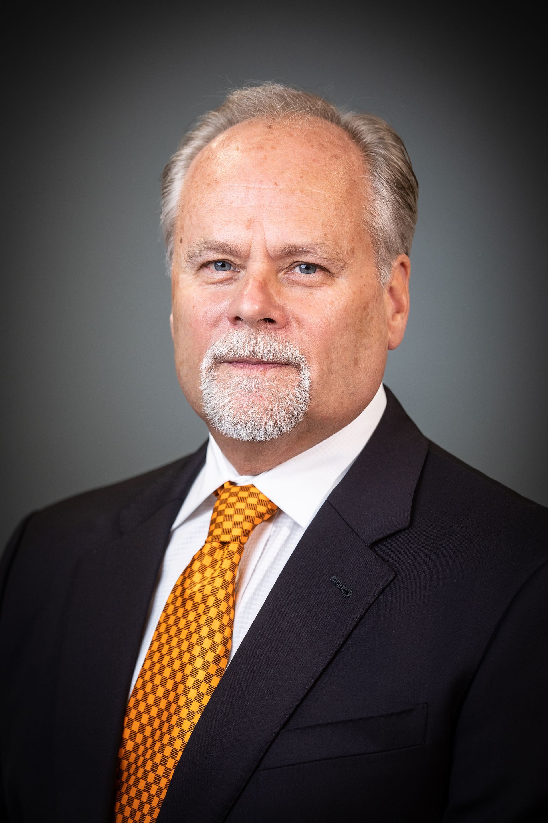 Studio portrait of a man in a dark suit, white shirt, and orange tie against a gray background