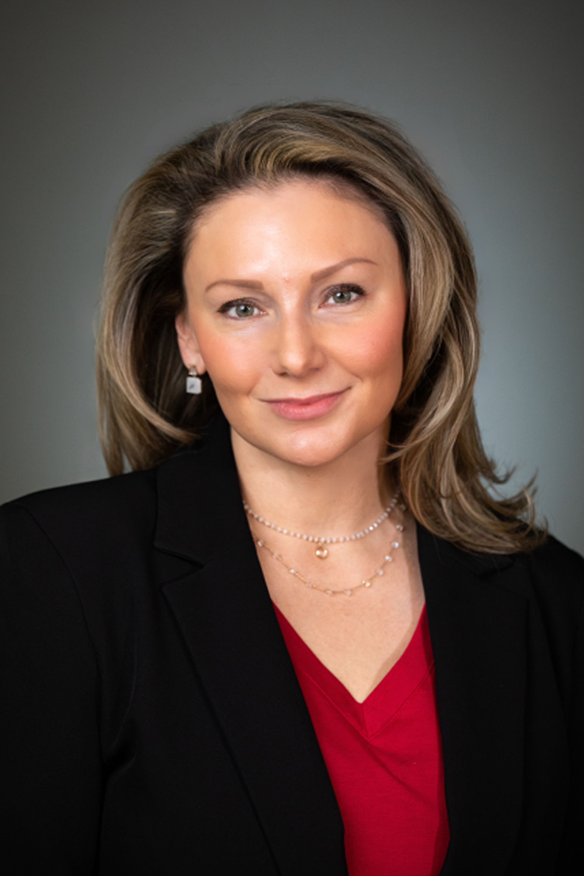 Professional headshot of a person in a black blazer and red top against a gray background