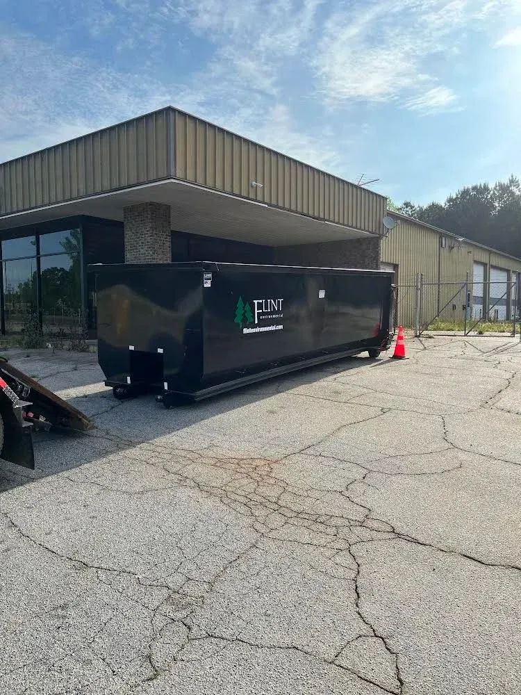 Black dumpster in front of a building with trees in background; sunny day.