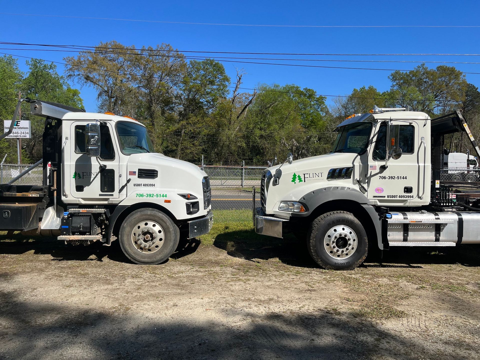 Two white dump trucks face each other outdoors with trees in the background under a blue sky.